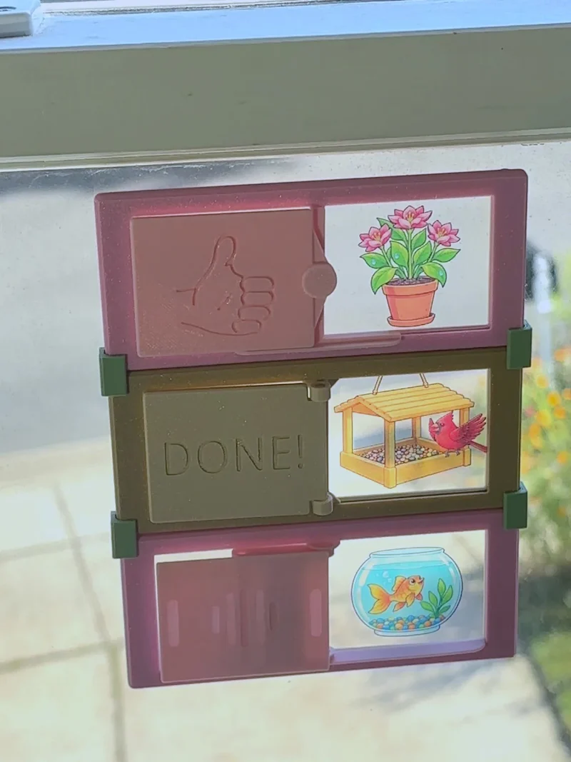 Three DoDones task frames clipped together on a window. The top cover is slid aside to show a potted plant card, the middle one is closed and reads DONE!, and the bottom shows a goldfish bowl card.